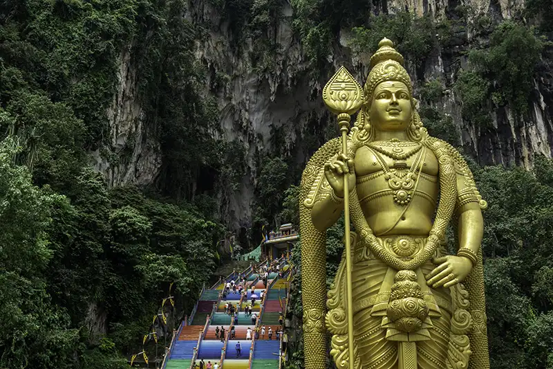 Large golden statue of Lord Murugan in front of the colorful steps leading up to Batu Caves, surrounded by lush green vegetation.