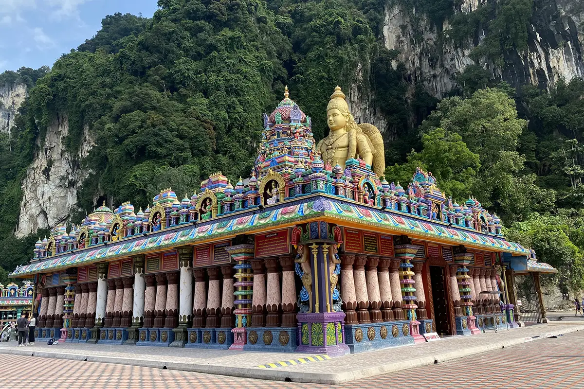 Colorful Hindu temple with intricate carvings and the Golden Lord Murugan statue at Batu Caves, Kuala Lumpur, surrounded by limestone hills and lush greenery.