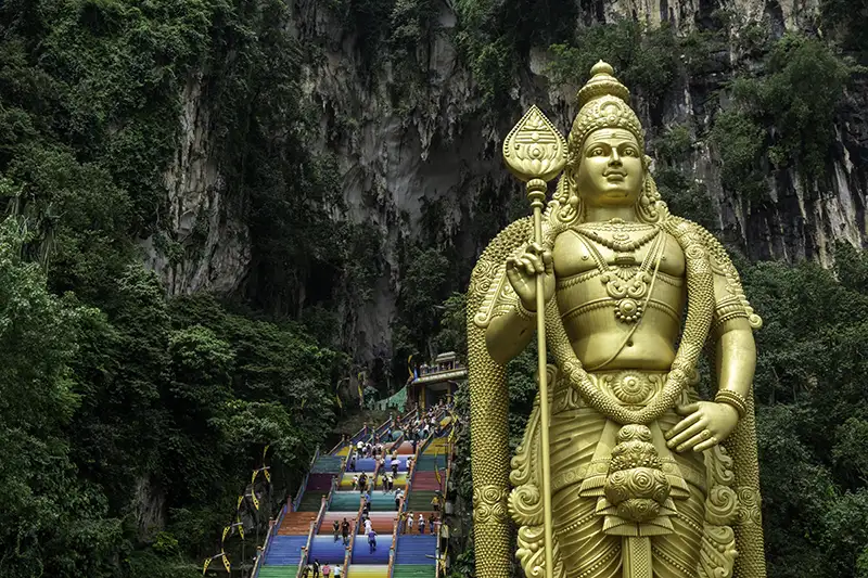 The Golden Lord Murugan statue at Batu Caves in Kuala Lumpur, with the colorful 272 steps and limestone cliffs in the background.