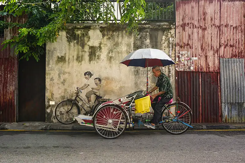 Trishaw rider on a street in George Town, Penang, with iconic street art in the background, a popular attraction for short trips in Malaysia.