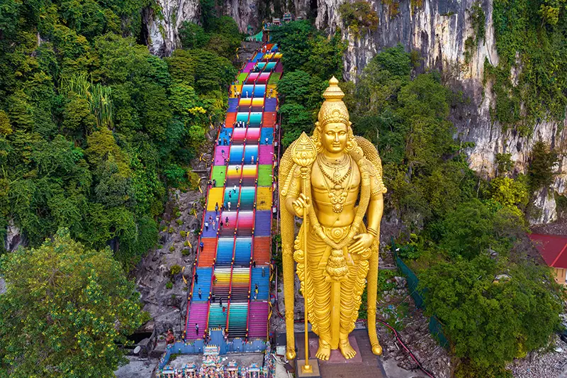 Aerial view of the 272-step Batu Caves staircase and the towering golden statue of Lord Murugan, set against limestone cliffs and lush greenery.